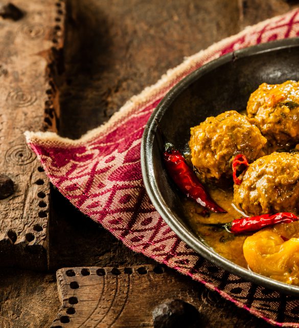 High Angle Close Up of Traditional Tajine Berber Dish of Meatballs Smothered in Spicy Yellow Curry Sauce with Hot Red Peppers and Served in Shallow Bowl on Cloth Napkin and Rustic Wooden Table