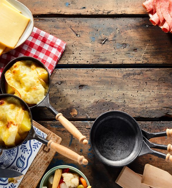 Traditional regional Swiss cuisine with melted raclette cheese over boiled potatoes served with cold meats, rustic border of the ingredients and skillets with copy space